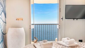 Een hotelkamer met een bed, een stoel, een televisie en een balkon met uitzicht op zee.