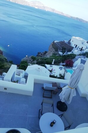 Una terraza con mobiliario blanco, una mesa y una sombrilla, con vistas a la costa y al mar, con acantilados rocosos.