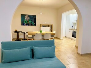 Ein modernes Wohnzimmer mit einem blauen Sofa, einem Essbereich mit einem Holztisch und Stühlen sowie einer Küche im Hintergrund.