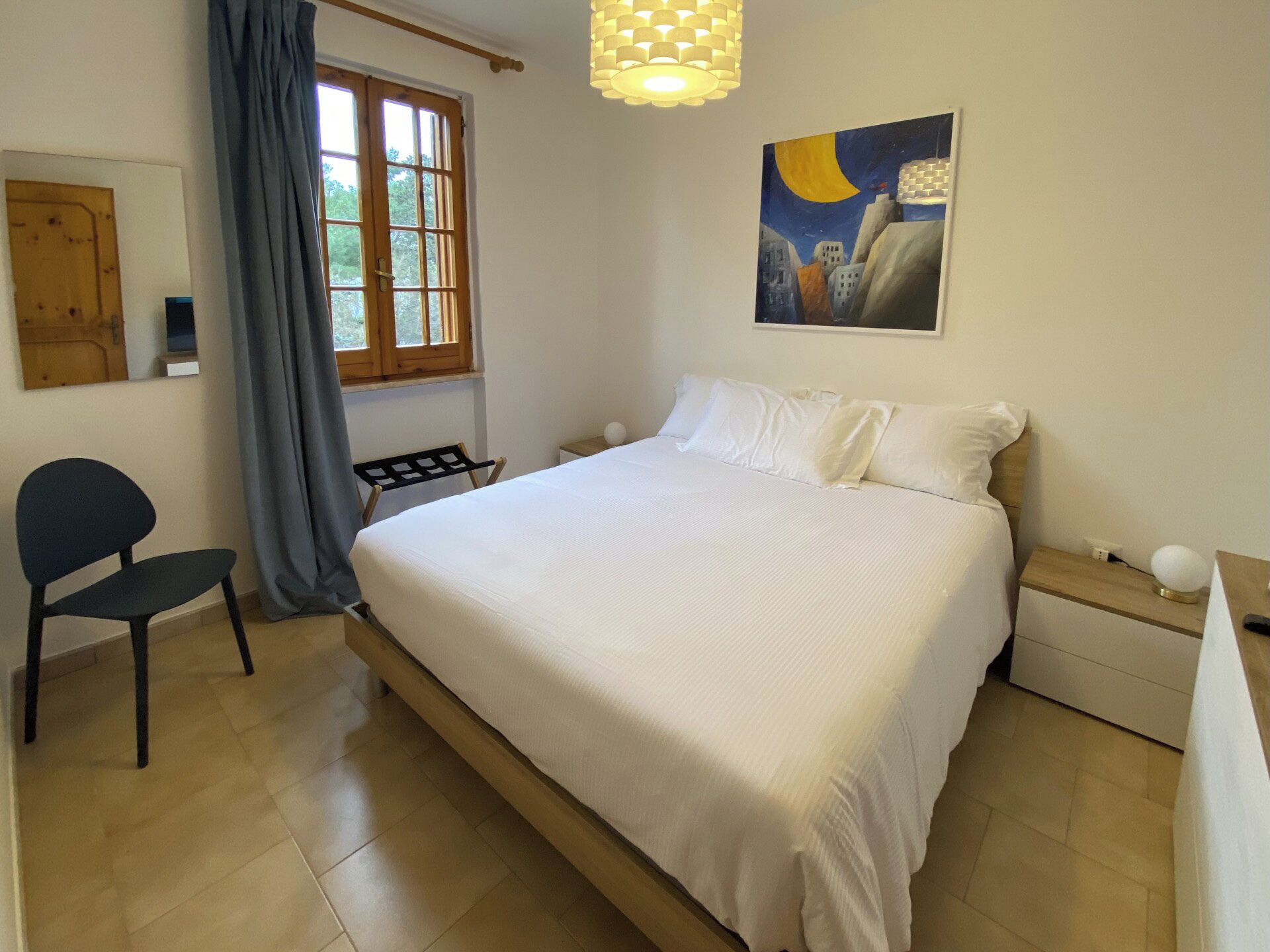 A bedroom with a large bed, a chair, a nightstand, a painting, and a window with curtains.