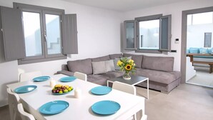 Una sala de estar moderna con una mesa de comedor blanca dispuesta para cuatro personas, sofás grises y un jarrón con flores.