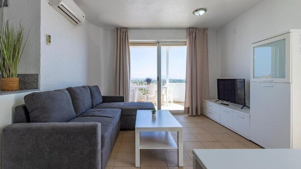 Una sala de estar moderna con un sofá gris, una mesa de centro blanca, un televisor sobre un mueble blanco y un balcón con vistas.