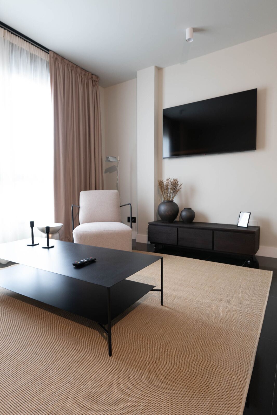 A modern living room with a flat-screen TV mounted on the wall, a black coffee table, a beige sofa, and a dark wooden cabinet with decorative items.