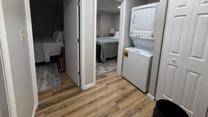 Uma lavanderia com máquina de lavar e secadora, uma cama com colcha verde e um tapete pequeno no chão.