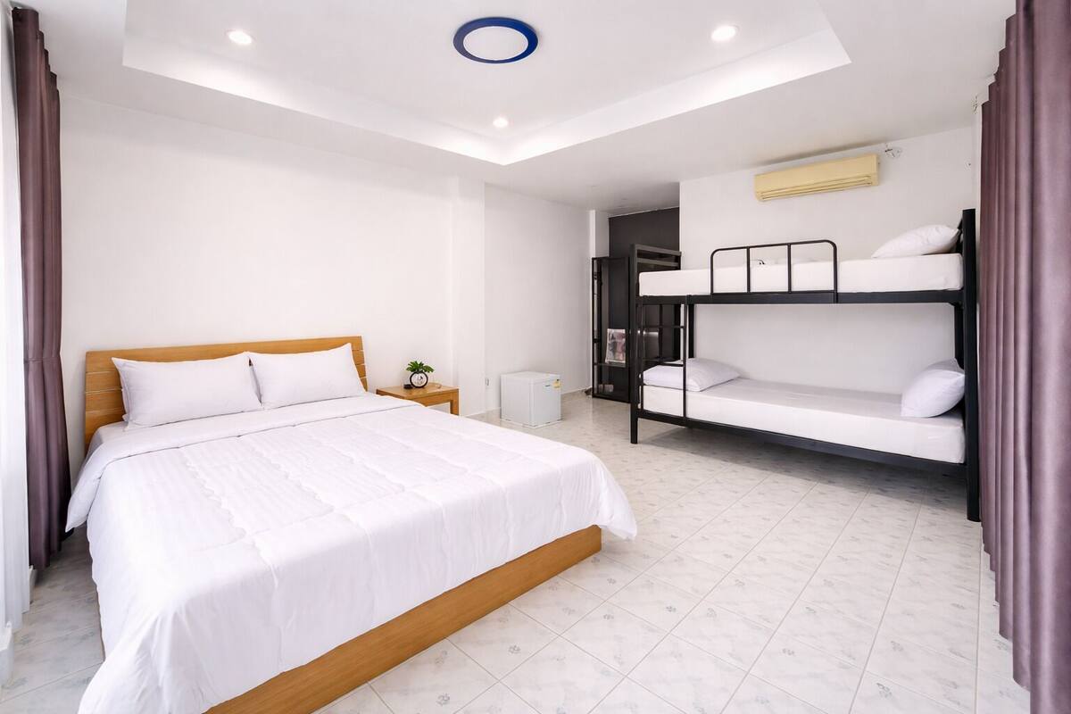 A hotel room with a single bed, a bunk bed, a small bedside table with a clock, and a white ceiling with a circular light fixture.