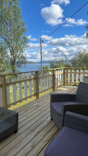 Une terrasse en bois avec des meubles en osier, une clôture blanche en bois et une vue sur un lac et des montagnes.