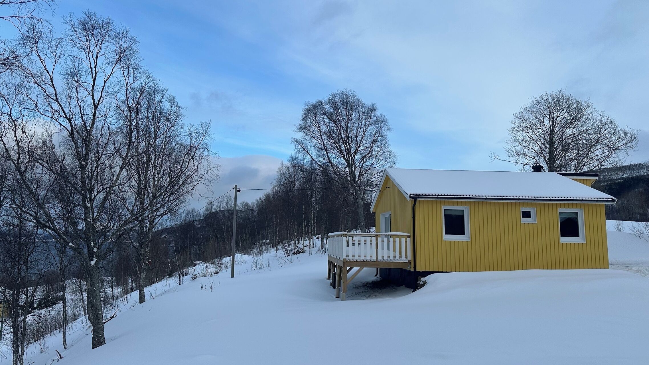 En gul hytte med en hvid veranda, omgivet af sneklædte træer og en klar himmel.
