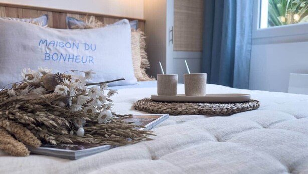 A neatly made bed with a white pillow labeled "MAISON DU BONHEUR," a woven tray with two cups, and a magazine on the bed. - House between forest & beach on foot • Nordic bath (Jard-sur-Mer)