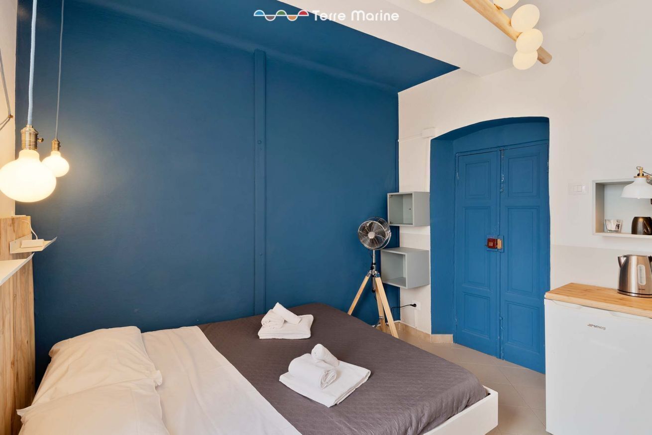 A bedroom with a blue accent wall, a bed with white linens, a small desk with a kettle, and a fan.