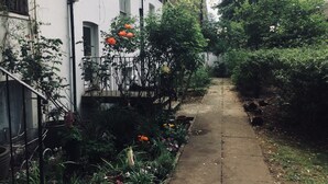 A narrow pathway flanked by lush greenery and a white building with multiple windows. - Boutique Bijoux Garden Flat in Vibrant Oval (London)