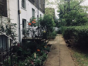 A narrow pathway flanked by lush greenery and a white building with multiple windows. - Boutique Bijoux Garden Flat in Vibrant Oval (London)