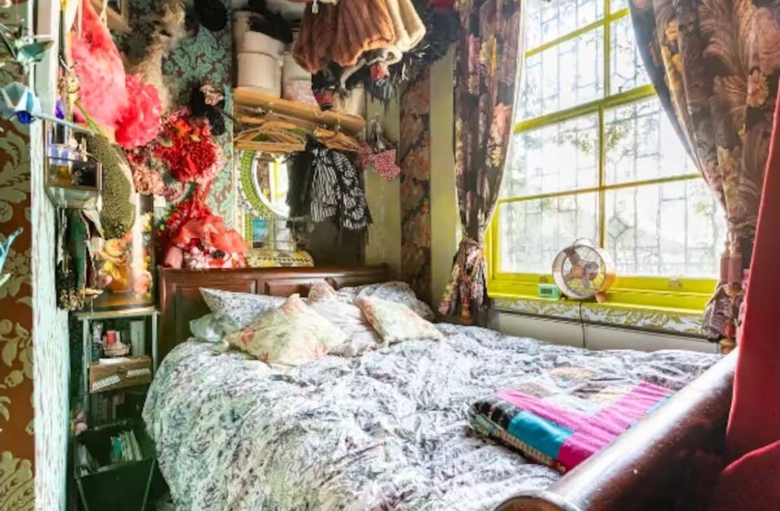 Ein Zimmer mit einem Bett, einem Vorhangfenster, einem Ventilator und verschiedenen Gegenständen auf Regalen und dem Bett.