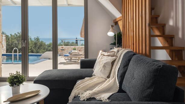 A modern living room with a grey sofa, a white table, and a view of a pool and outdoor seating area. - Villa Kamaria - 2 Bedrooms with Private Pool and Sea View (Zakynthos)