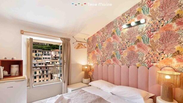 A hotel room with a bed, a window with a view of buildings, a patterned wall, and a bedside table with a lamp.