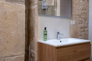 Un baño con lavamanos blanco, mueble de madera, pared de piedra y un espejo sobre el lavamanos.