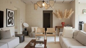 Een moderne woonkamer met een bank, twee fauteuils, een salontafel en een eettafel met stoelen. Er hangt een lamp en er is een ingelijst kunstwerk aan de muur.