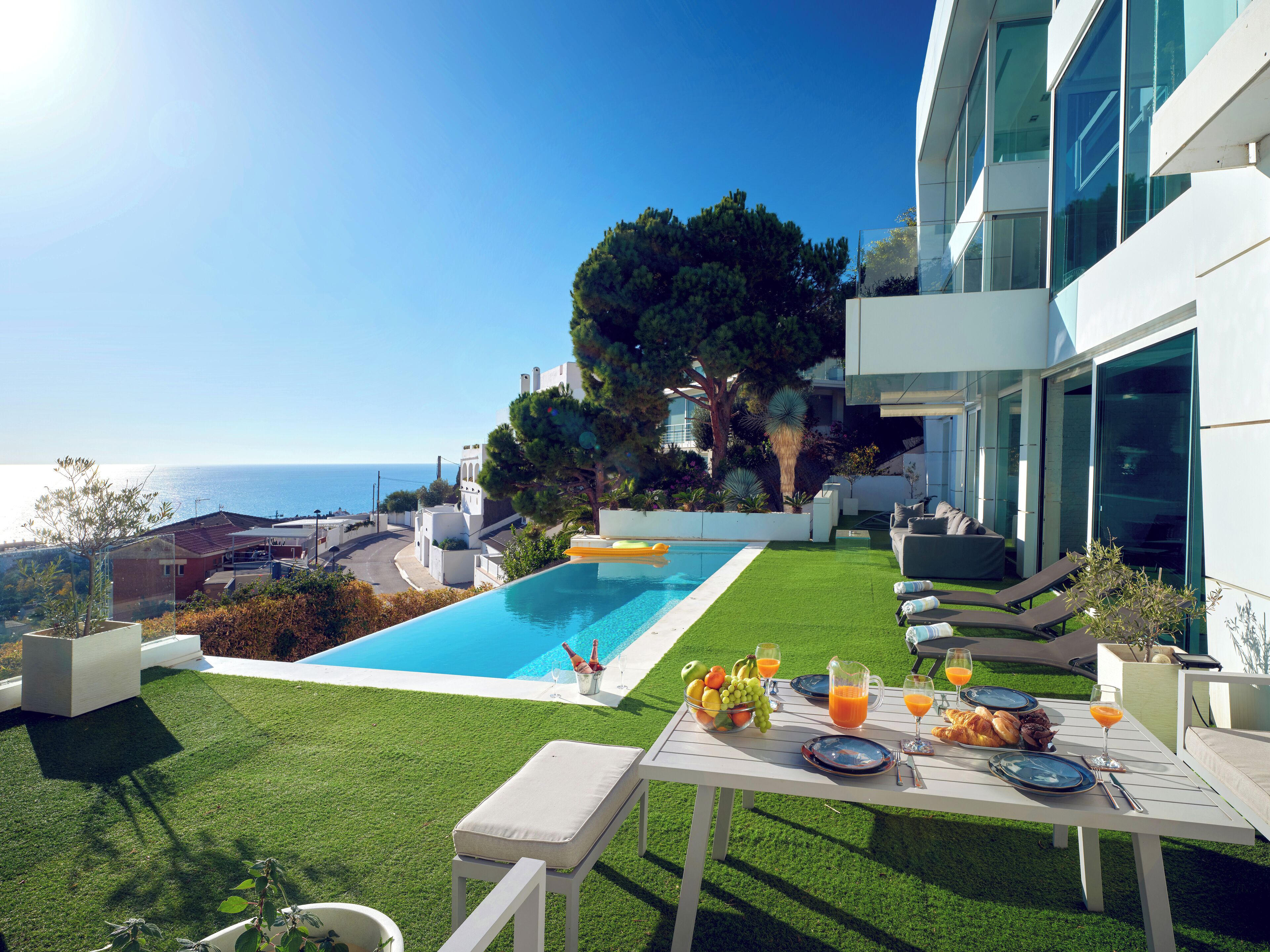 A modern outdoor pool area with a dining table set for breakfast, overlooking a scenic ocean view.