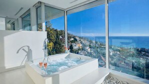 A modern bathroom with a large bathtub, a glass shower enclosure, and a view of a coastal town. - Villa in Sitges |Pool|Jacuzzi|BBQ|Views|AC|12P (Sitges)