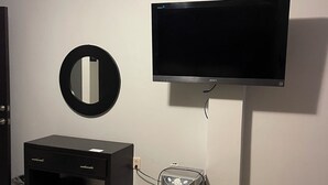 Una habitación con un televisor negro en la pared, un escritorio pequeño con cajones, un cubo de basura y un calentador.
