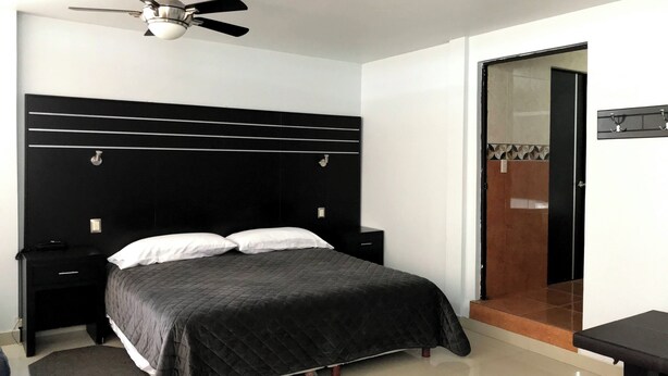 Un dormitorio con cabecera negra, cama con colcha acolchada oscura, mesita de noche y puerta con espejo y perchero para toallas.