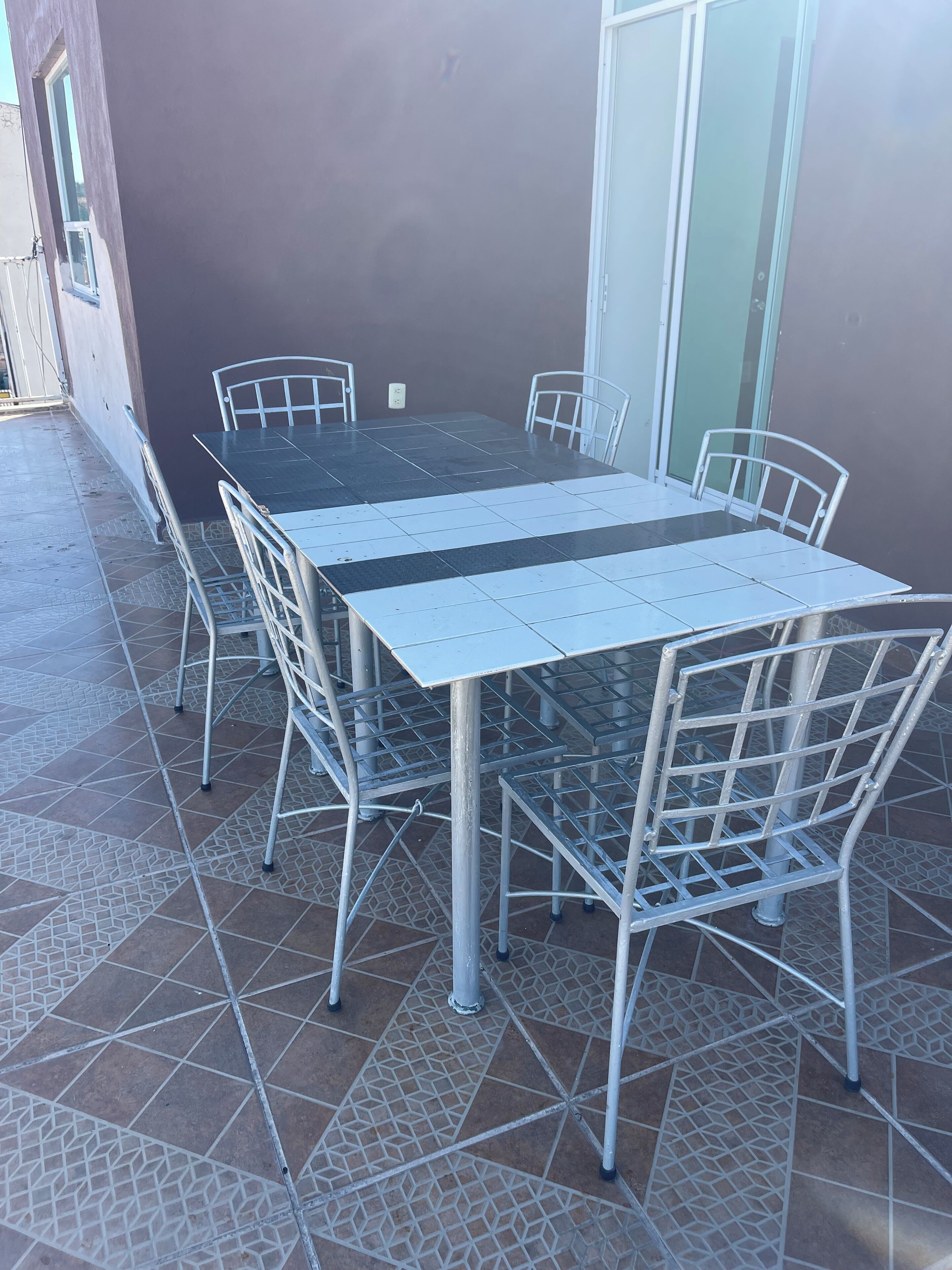 A rectangular outdoor dining area with a metal table and chairs, surrounded by a tiled floor with a geometric pattern.
