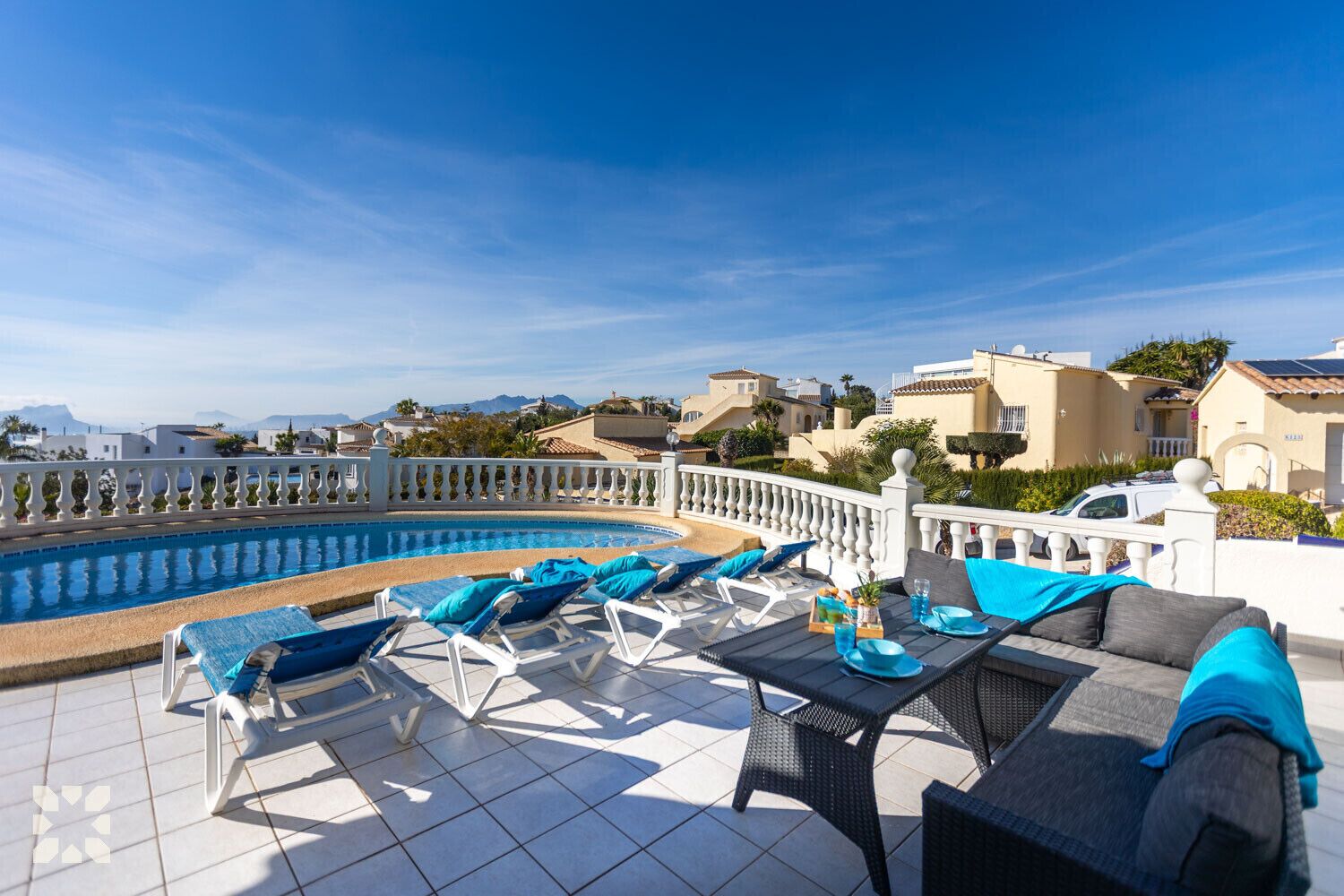 A rooftop pool area with lounge chairs and a table, surrounded by a white railing and overlooking a residential area with houses and mountains in the background.