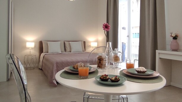 Ein modernes Hotelzimmer mit einem runden Esstisch, transparenten Stühlen, einem Bett mit rosa Bettdecke, einem Nachttisch mit Lampe und einem Fenster mit Vorhängen.