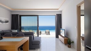 A modern living room with a large window overlooking the ocean, a grey sofa, a wooden dining table, and a flat-screen TV. - Falasarna Village  (Kissamos)