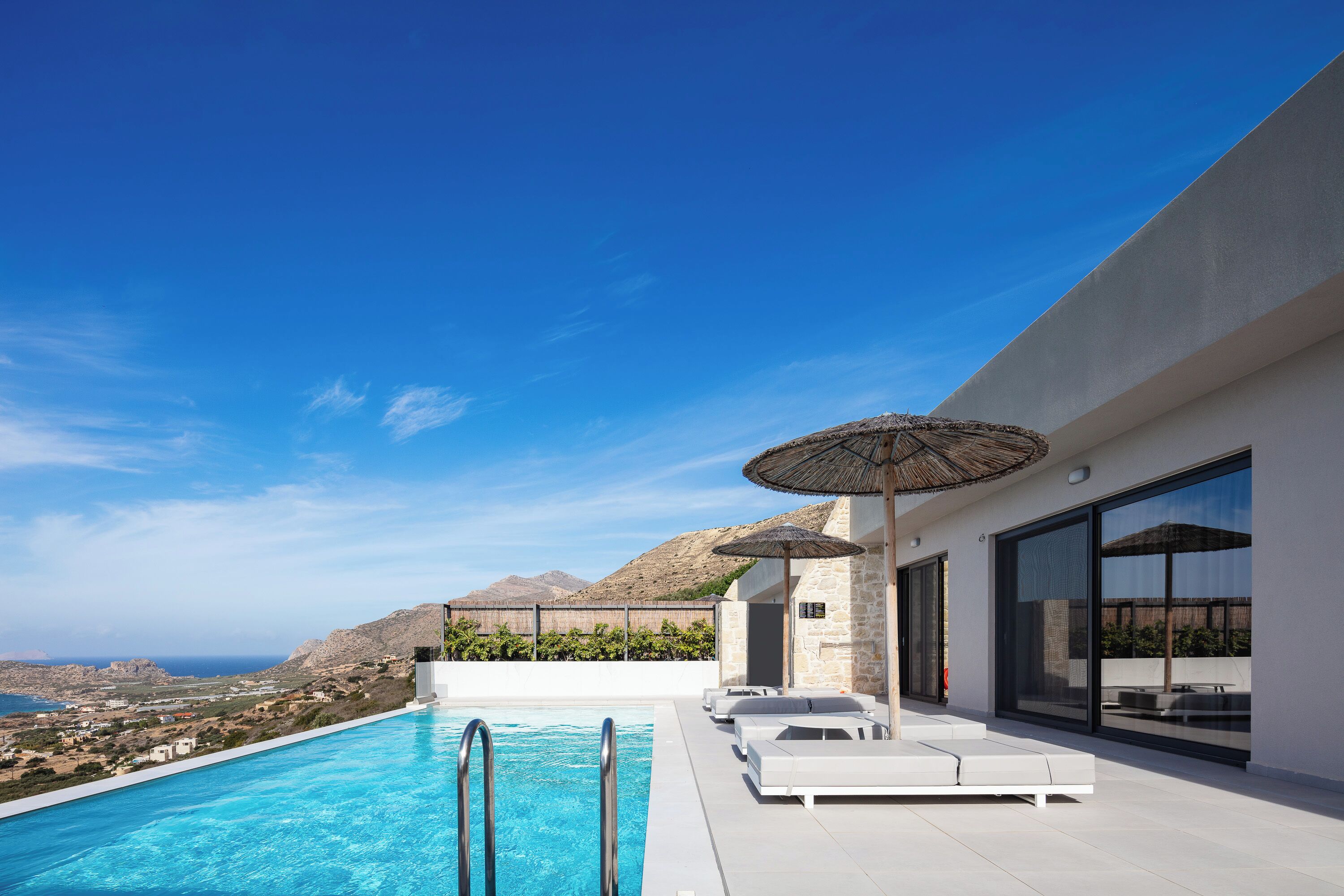 A modern outdoor pool area with clear blue water, white lounge chairs, and umbrellas, overlooking a scenic coastal landscape.