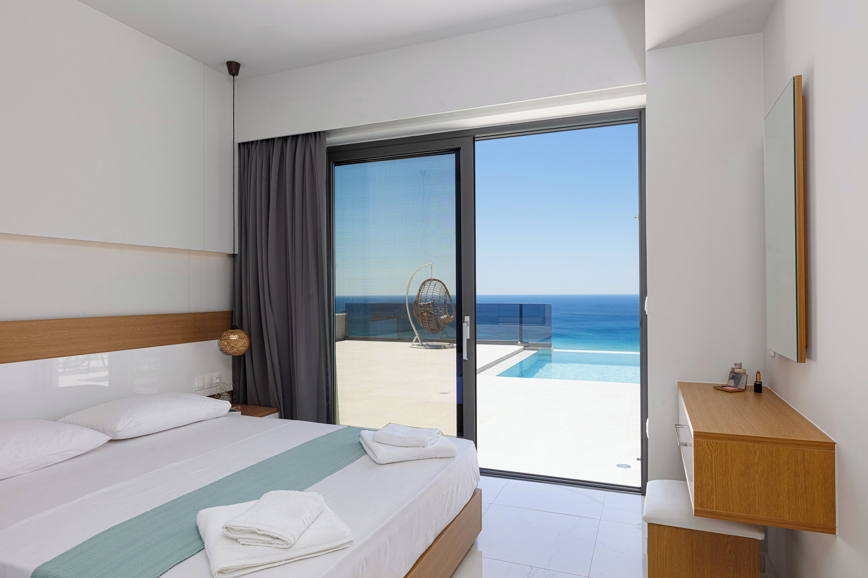 Ein Hotelzimmer mit Bett, Nachttisch, Holzbeistelltisch, einem großen Fenster mit Meerblick und einer Glastür zum Balkon.