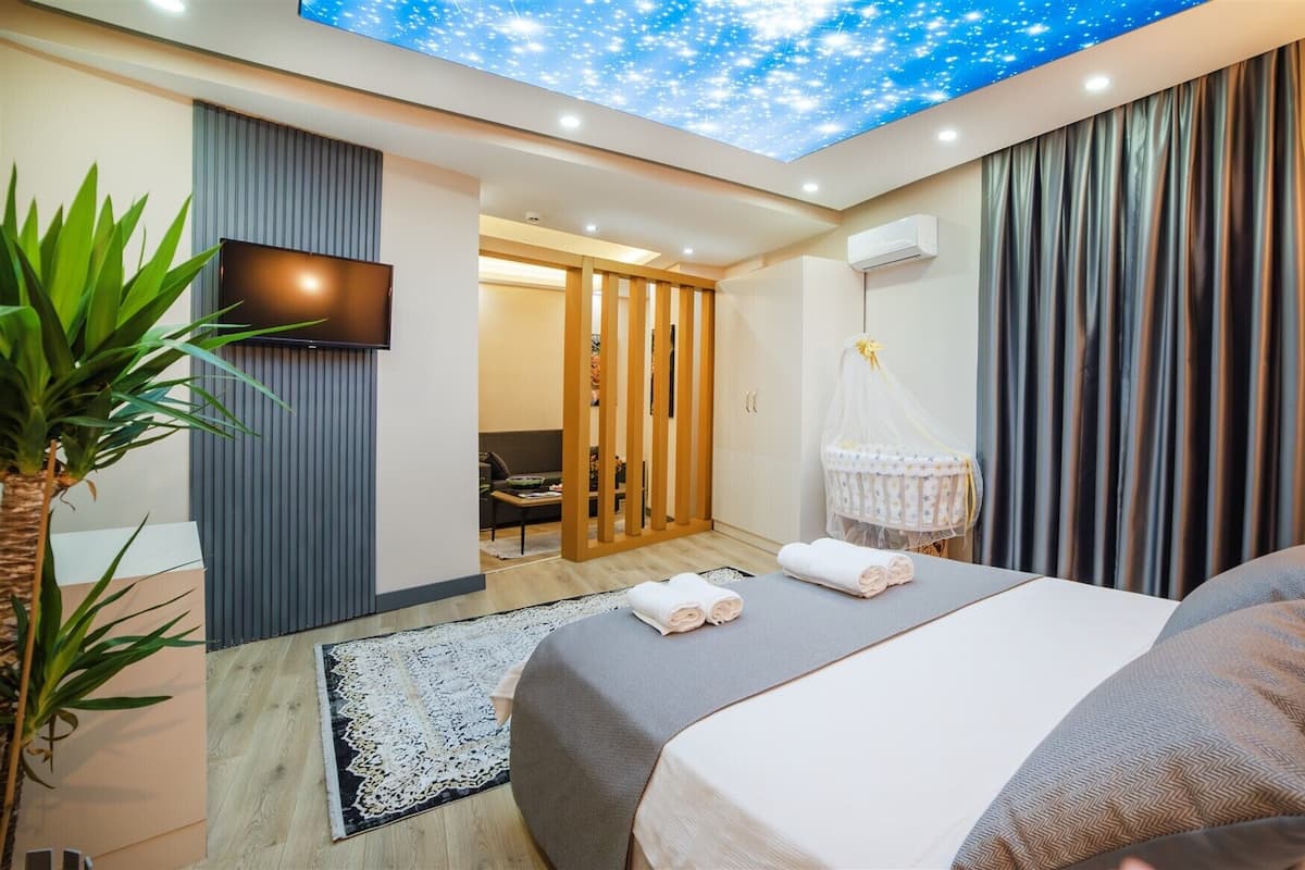 Una habitación de hotel moderna con cama, televisión, proyector en el techo, una planta y vista a la habitación contigua.