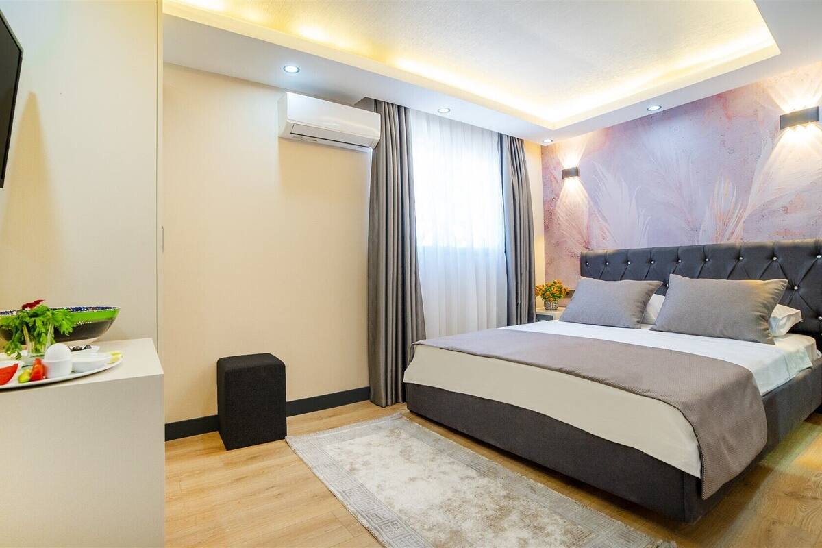 Una habitación de hotel moderna con una cama grande, una mesita de noche con un frutero, un televisor de pantalla plana y un aire acondicionado instalado en la pared.
