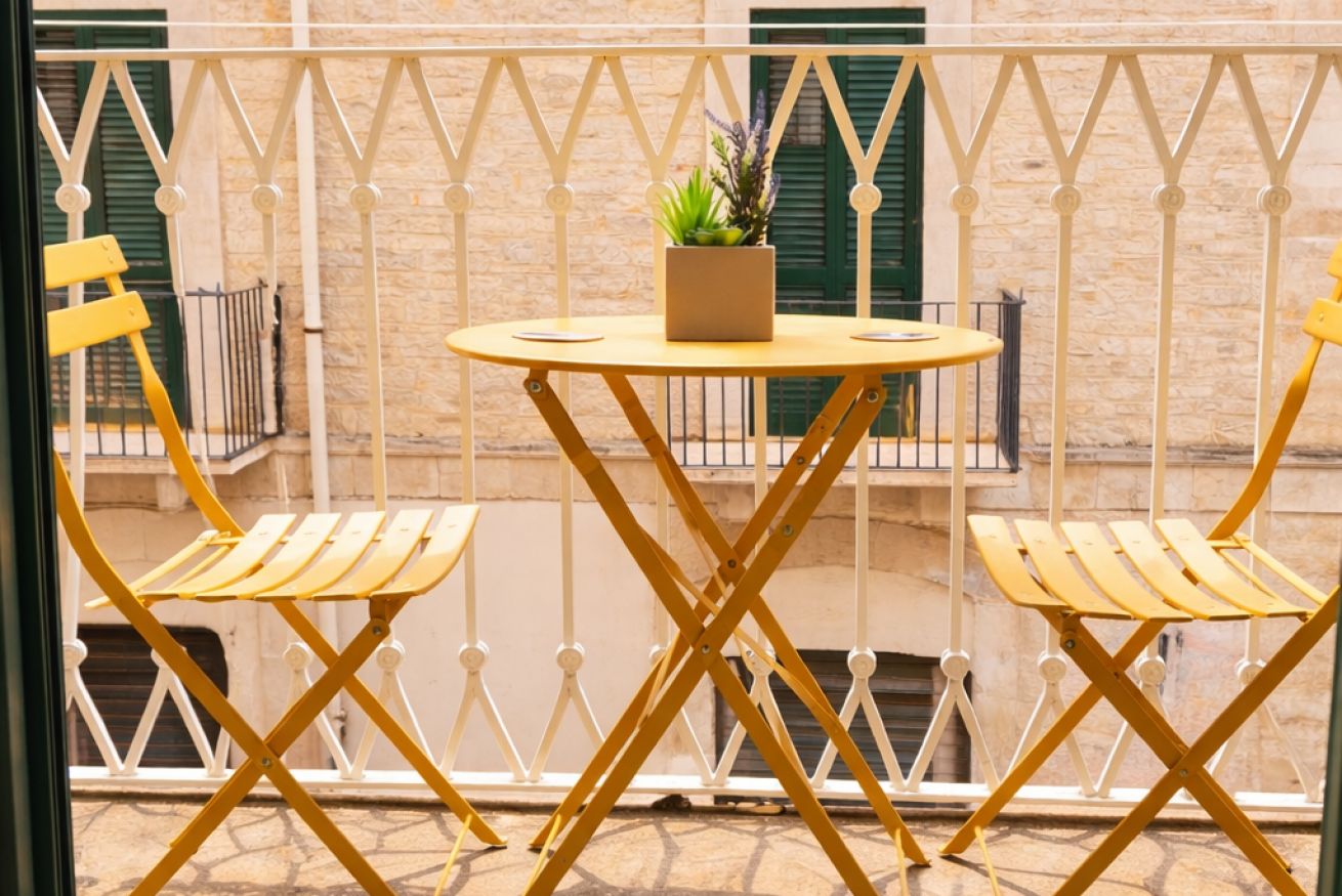Een balkon met een ronde tafel en twee stoelen, een bloempot en uitzicht op een gebouw met stenen muren en groene luiken.