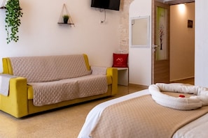 Una stanza con un divano giallo, un comodino, un letto bianco con un cuscino bianco e un cuscino rosso, una TV a parete e una porta con decorazioni verdi.