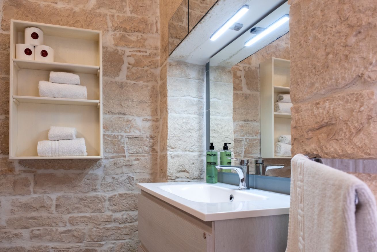 Un baño con paredes de piedra, un lavabo blanco y estantes con toallas y artículos de aseo.