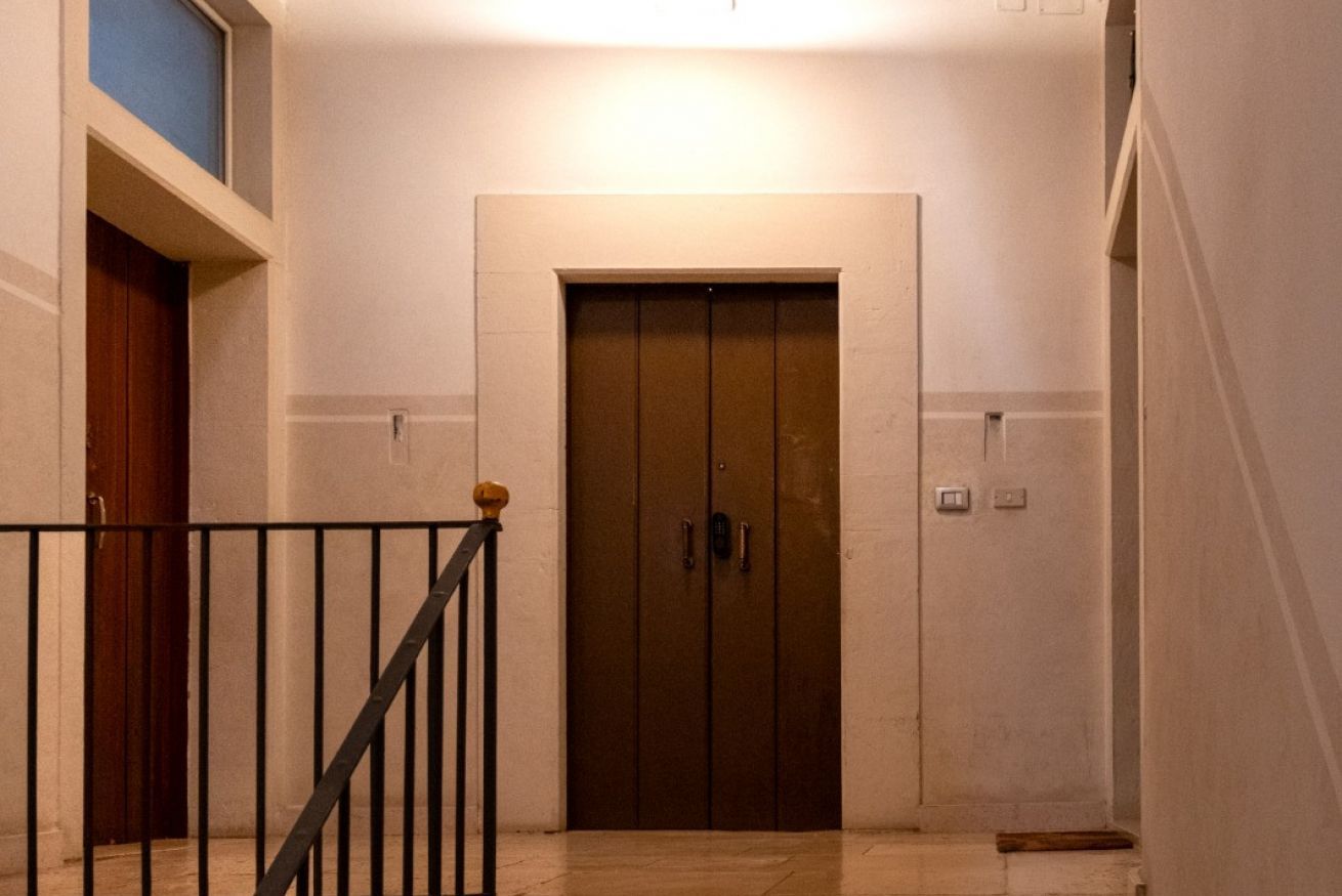 Un couloir avec une porte d’ascenseur fermée, une porte en bois sur la gauche et une rambarde noire.