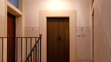 Un couloir avec une porte d’ascenseur fermée, une porte en bois sur la gauche et une rambarde noire.