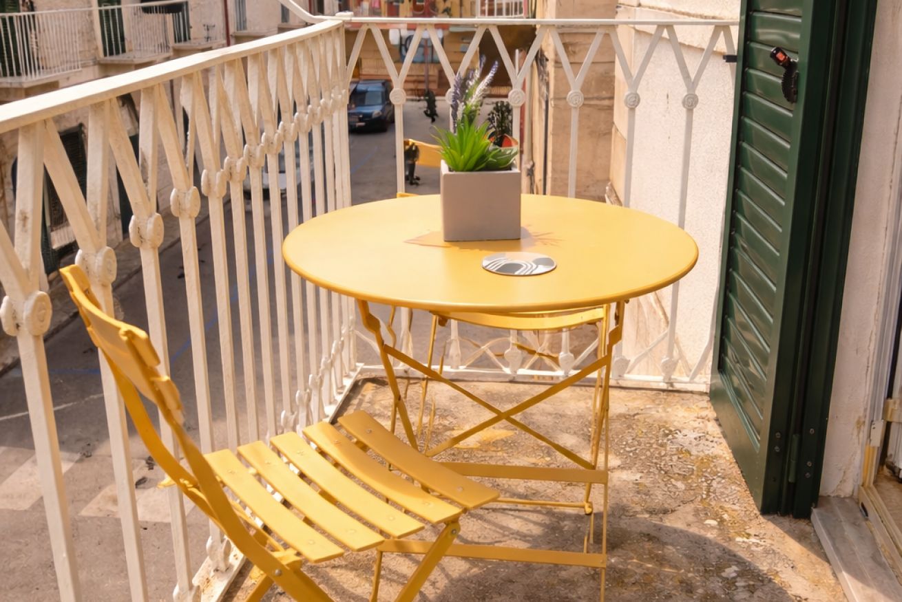 Een gele buitentafel met een potplant, een stoel en een witte leuning.