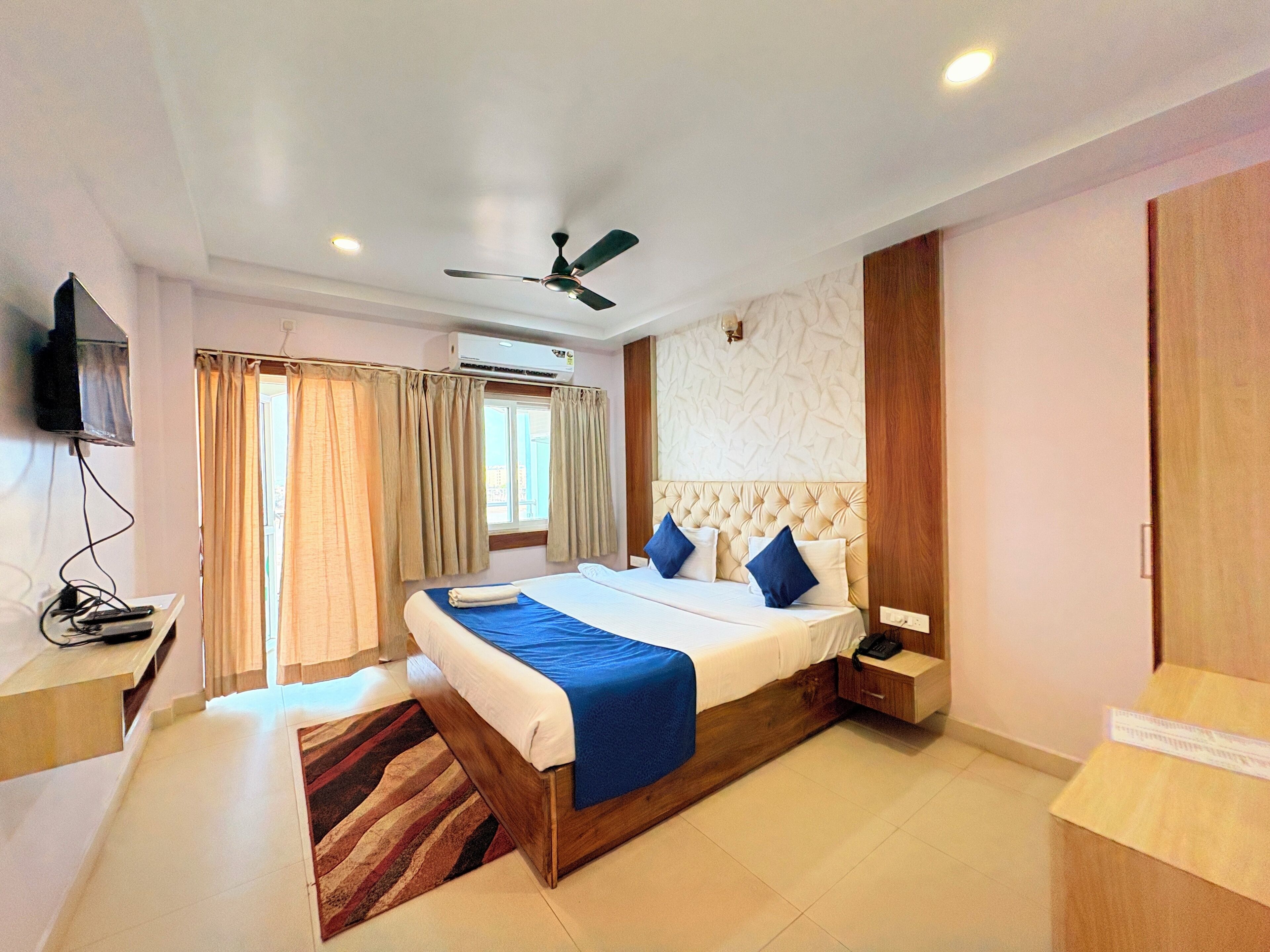 A hotel room with a bed, a ceiling fan, an air conditioning unit, a TV mounted on the wall, and a window with curtains.