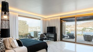 A modern hotel room with a bed, a TV, a seating area with a table, and a balcony with a view of the sea and buildings. - Villa Elettra close to the sea - My Rental Homes (Mykonos)