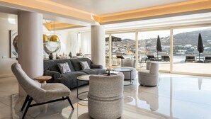 A spacious living area with a sofa, armchairs, and ottomans, a round coffee table, and a view of the sea through large windows. - Villa Elettra close to the sea - My Rental Homes (Mykonos)