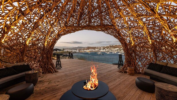 An outdoor seating area with a fire feature, wicker furniture, and a view of a coastal town. - Villa Elettra close to the sea - My Rental Homes (Mykonos)
