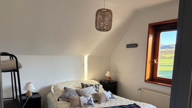 Een slaapkamer met een bed, een nachtkastje, een lamp, een hangende lamp, een raam met uitzicht op het landschap en een bord aan de muur.