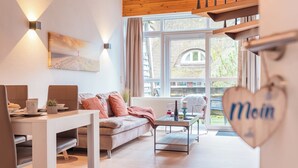 Ein modernes Wohnzimmer mit einer Couch, einem Esstisch und einem Couchtisch. Ein großes Fenster bietet Blick auf ein Gebäude und Bäume.