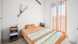 Un dormitorio con cama, una mesita de noche de madera, una estantería y una ventana con cortinas traslúcidas.