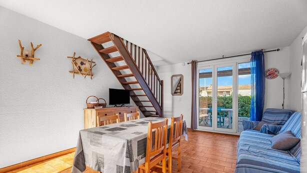 Un área de comedor con una mesa dispuesta para cuatro, una escalera de madera, un sofá y un televisor sobre un mueble de madera.