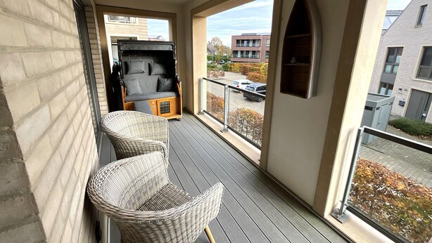 A balcony with wicker chairs, a sofa, and a view of a parking lot and buildings. - Hello and welcome to Nordseeliebe! (Dangast)