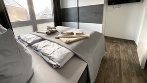 A modern bedroom with a large bed, a flat-screen TV mounted on the wall, and a window with curtains. - Moin and welcome to Apartment Küstenglück! (Dangast)