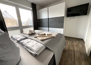 A modern bedroom with a large bed, a flat-screen TV mounted on the wall, and a window with curtains. - Moin and welcome to Apartment Küstenglück! (Dangast)