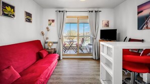 Una habitación con un sofá rojo, televisión, zona de comedor con sillas rojas y balcón con mesa y sillas.